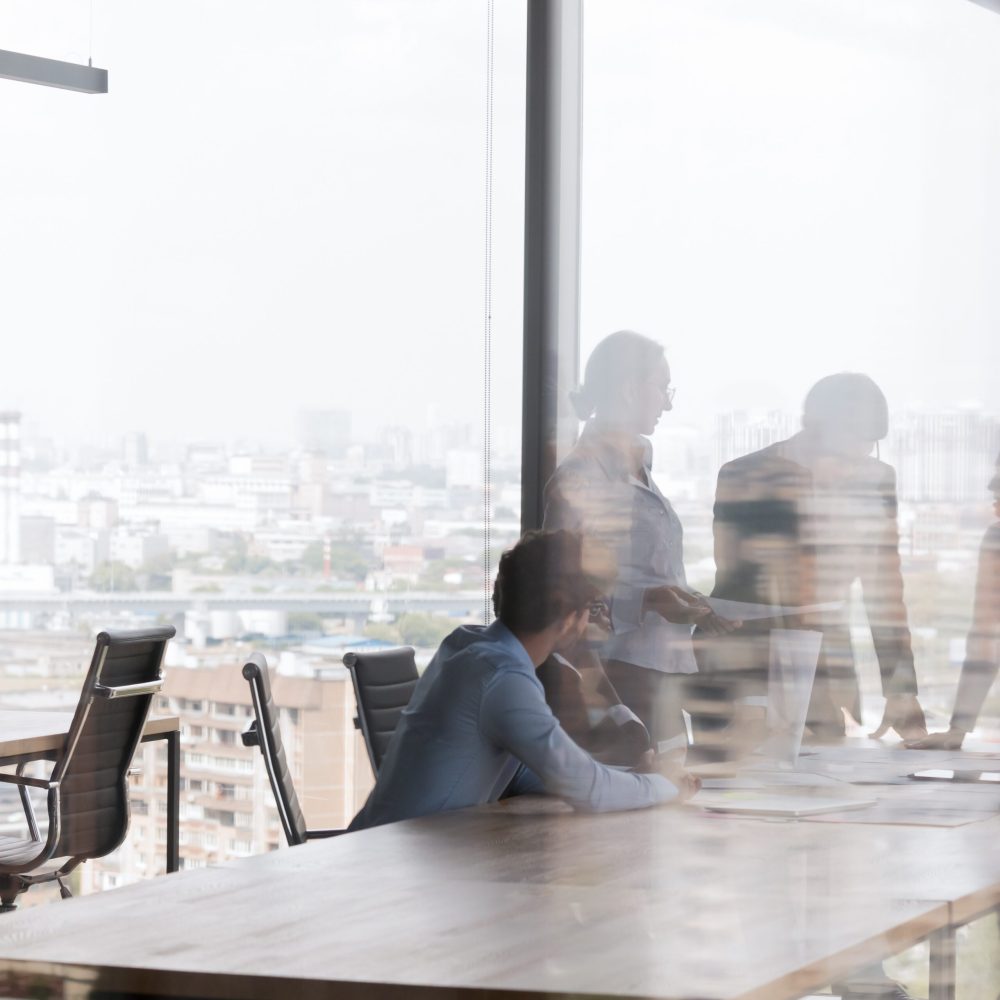 Business team meeting in contemporary business center, talking at office conference table, negotiating on project in modern interior with big panoramic window. Through glass view background